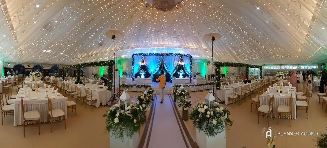 Wide view of a tented reception: a flower-lined aisle, dining tables and a lit stage