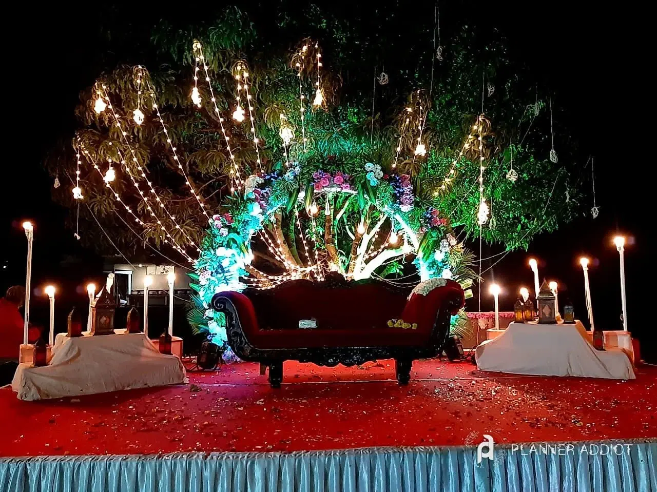 Night stage under a tree hung with lights, with a floral arch, a red sofa and tall candles
