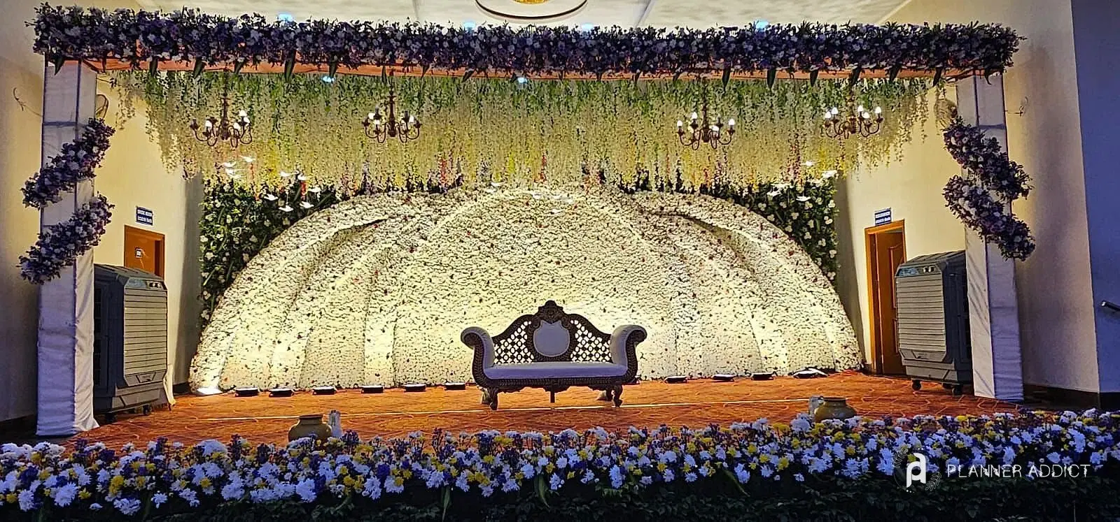 Reception stage with a white flower dome, hanging wisteria and a carved sofa
