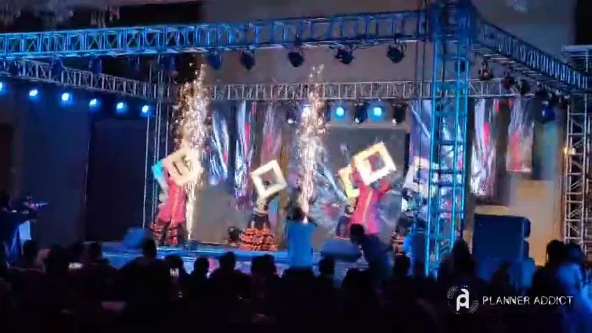 Performers on a truss stage with moving lights, LED panels and cold fireworks, in front of a seated audience