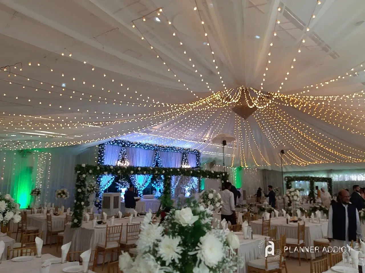 Reception under a white fabric ceiling strung with fairy lights, with a green-lit stage and dining tables